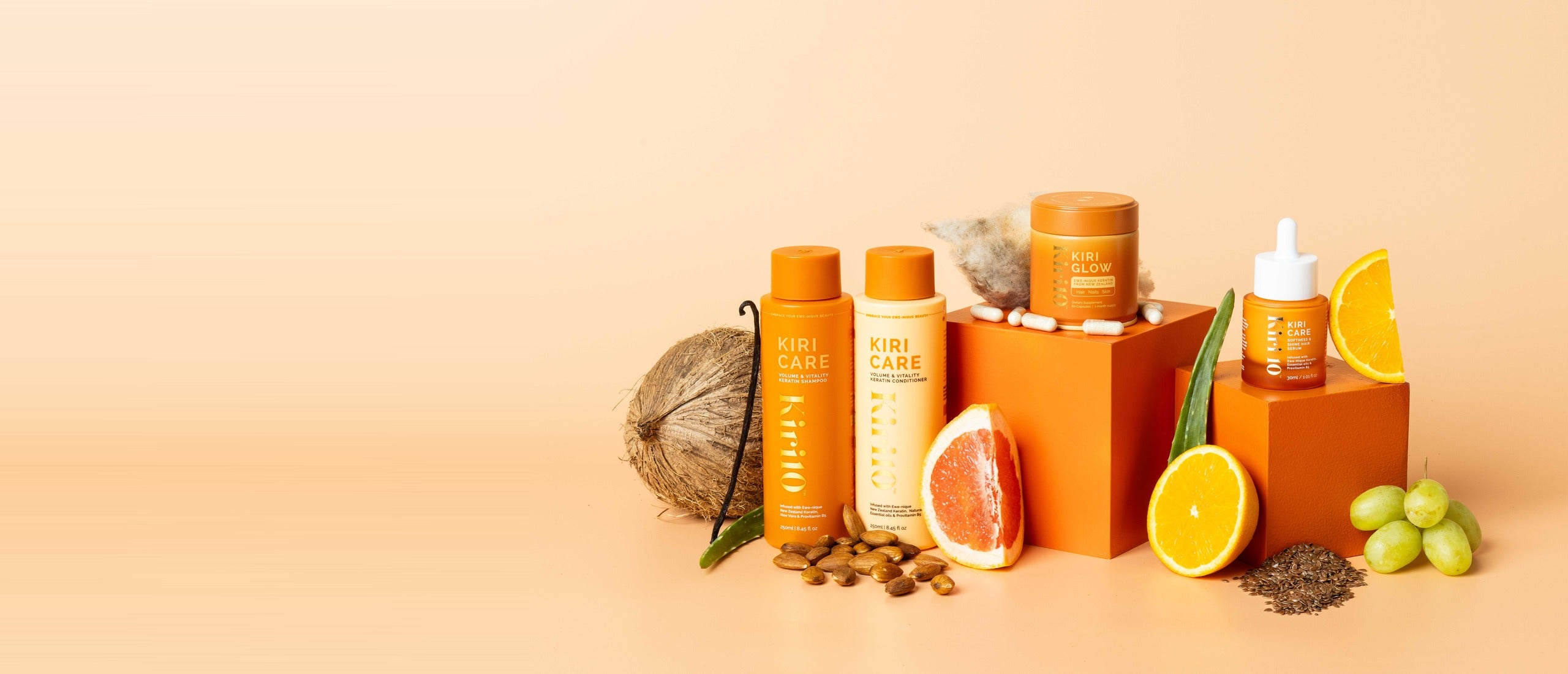 KiriGlow keratin capsules with the KiriCare shampoo, conditioner and hair serum on a peach backdrop, surrounded by sheep wool and the fruit and nut ingredients